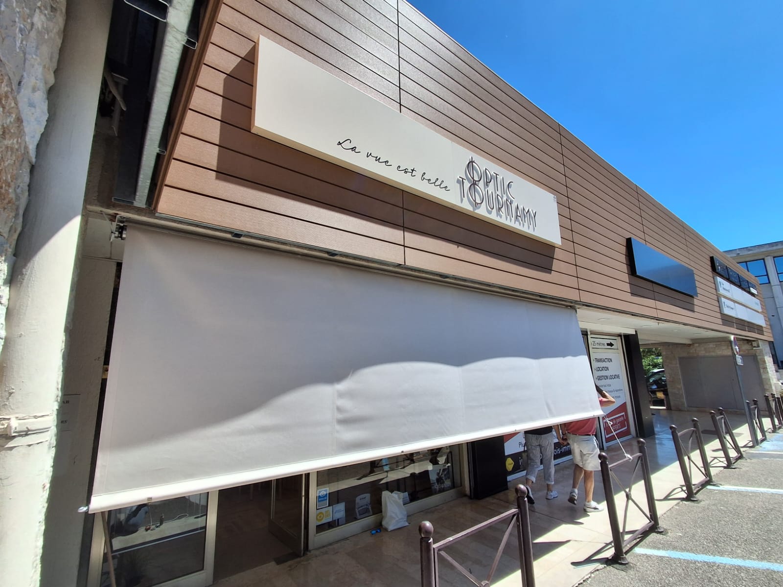 Store banne vertical blanc déployé sur la vitrine de l'opticien Optic Tournamy à Mougins, vue de face depuis le parking